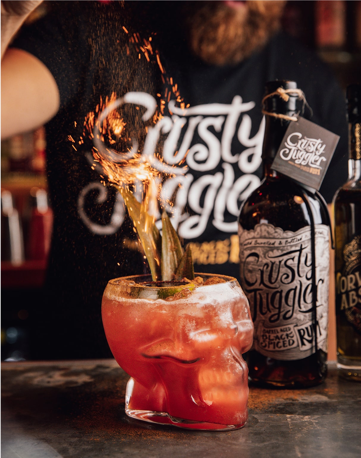 A bartender sprinkles sparks over a red cocktail in a skull-shaped glass, garnished with pineapple. Bottles of Gutsy Juggler Spiced Rum are visible, with a branded shirt in the background.