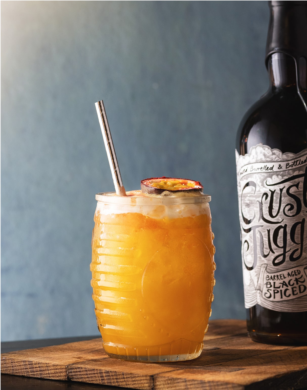 A honeycomb-patterned glass filled with an orange cocktail, garnished with a passion fruit slice and a straw, sits on a wooden surface next to a dark bottle labeled Crusty Juggler Black Spiced.