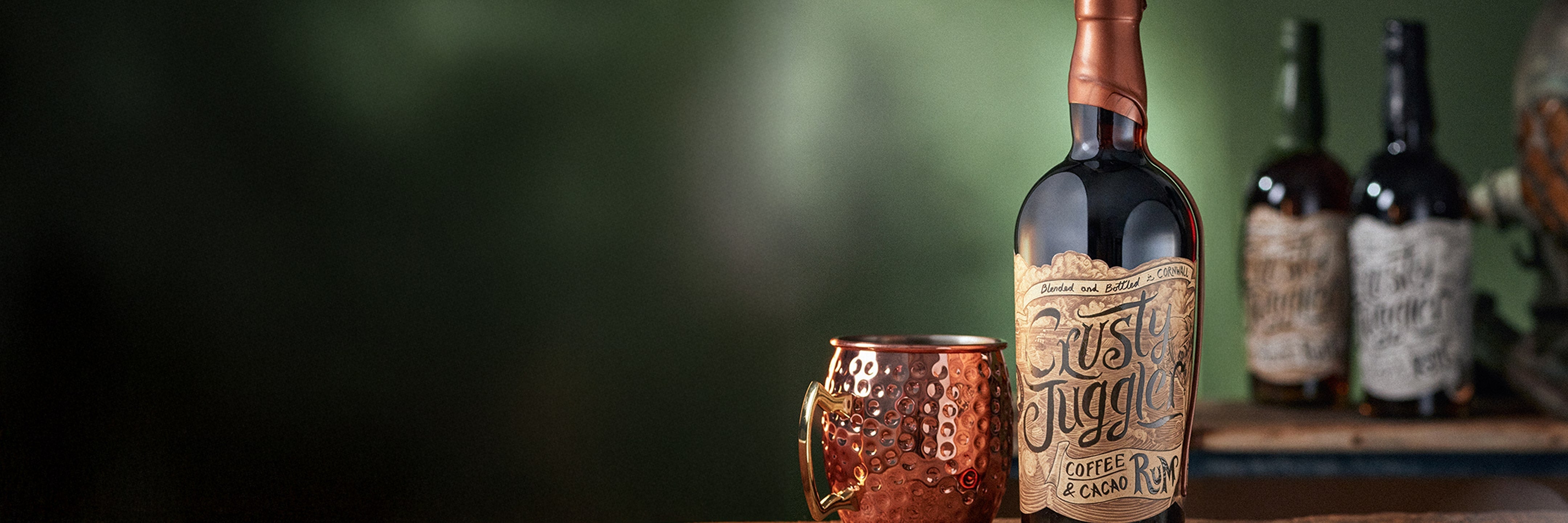 A bottle labeled Cask & Kettle sits next to a copper mug on a wooden surface, with two blurred bottles and a globe in the background against a green backdrop.