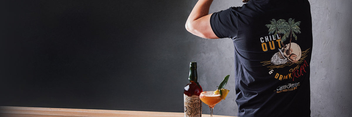 A person in a “Chill Out & Drink Rum” t-shirt stands at a wooden counter with a rum bottle and a tropical cocktail, facing away from the camera.