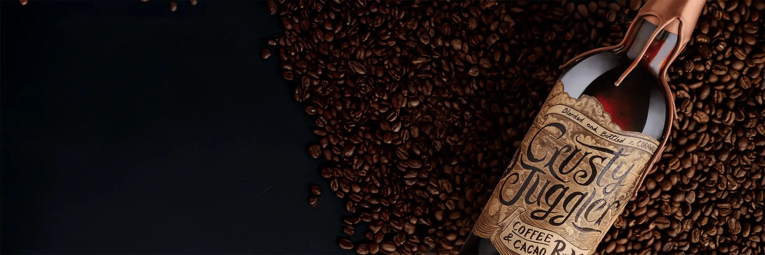 A bottle with a handwritten-style label reading “Cask Suggie Coffee Cacao” lies on its side next to a pile of coffee beans on a dark surface.