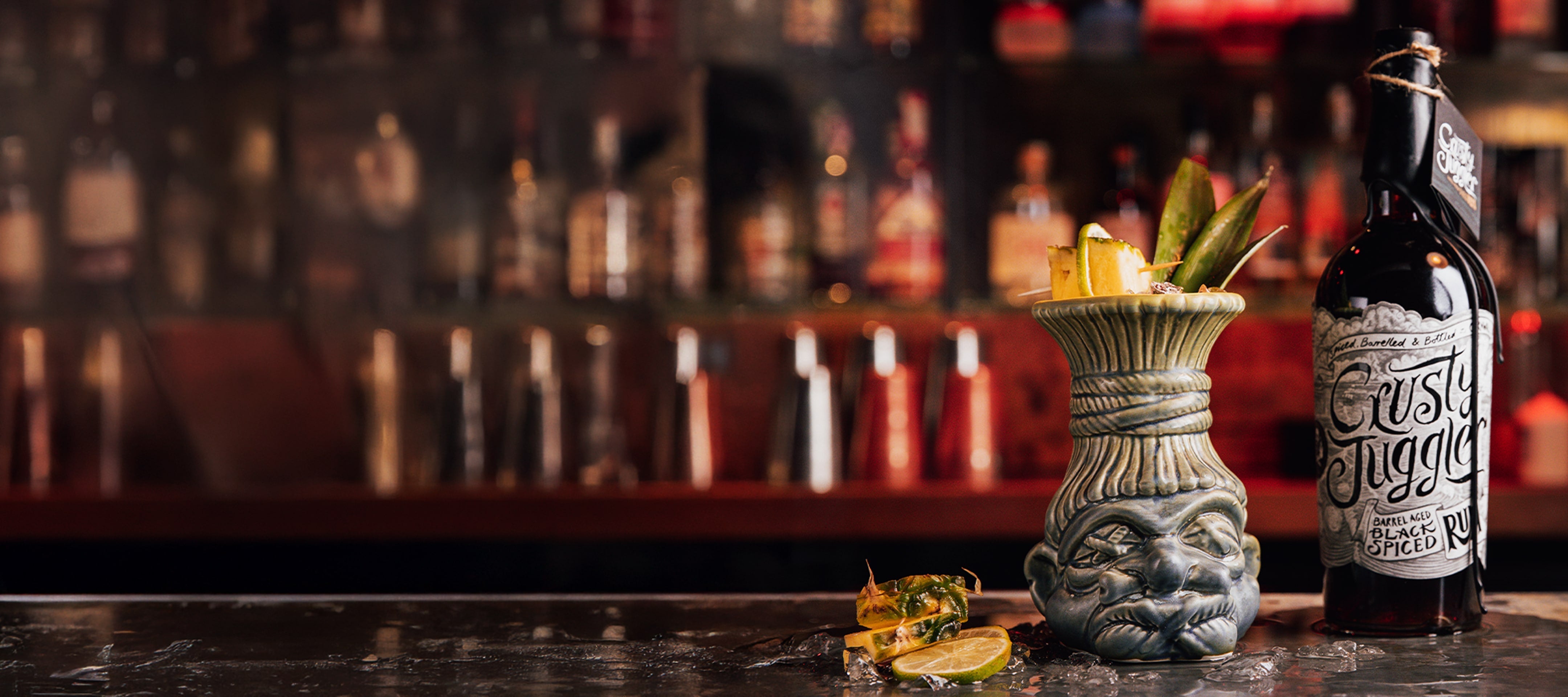 A tiki-style ceramic mug with a tropical drink, garnished with pineapple and leaves, sits on a bar next to a bottle of spiced rum. Lime wedges and pineapple pieces are on the counter.