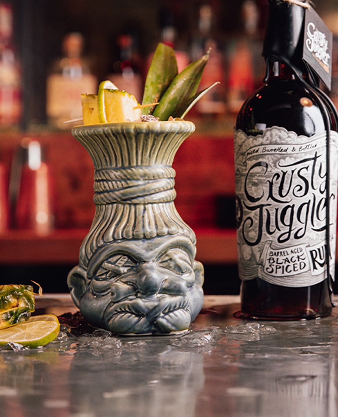 A tiki mug filled with a tropical cocktail garnished with pineapple and leaves sits on a bar next to a bottle of Crusty Juggler Black Spiced Rum; ice, lime, and pineapple are scattered on the counter.