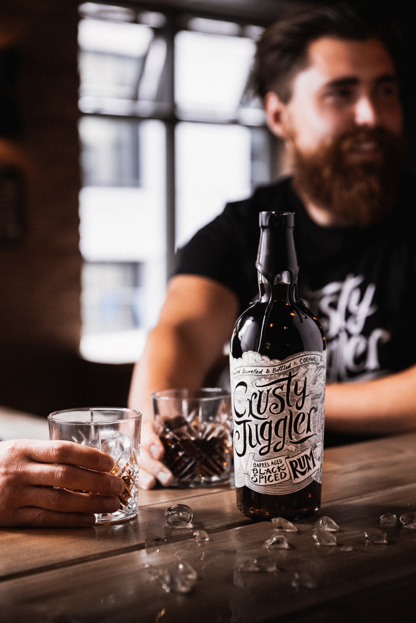 A man wearing a Crusty Classic Black Tee drinks at a wooden table. A 70cl bottle of Crusty Juggler Barrel Aged Black Spiced Rum stands on the table surrounded by crushed ice. A hand from a second person can also be seen holding a rocks glass.