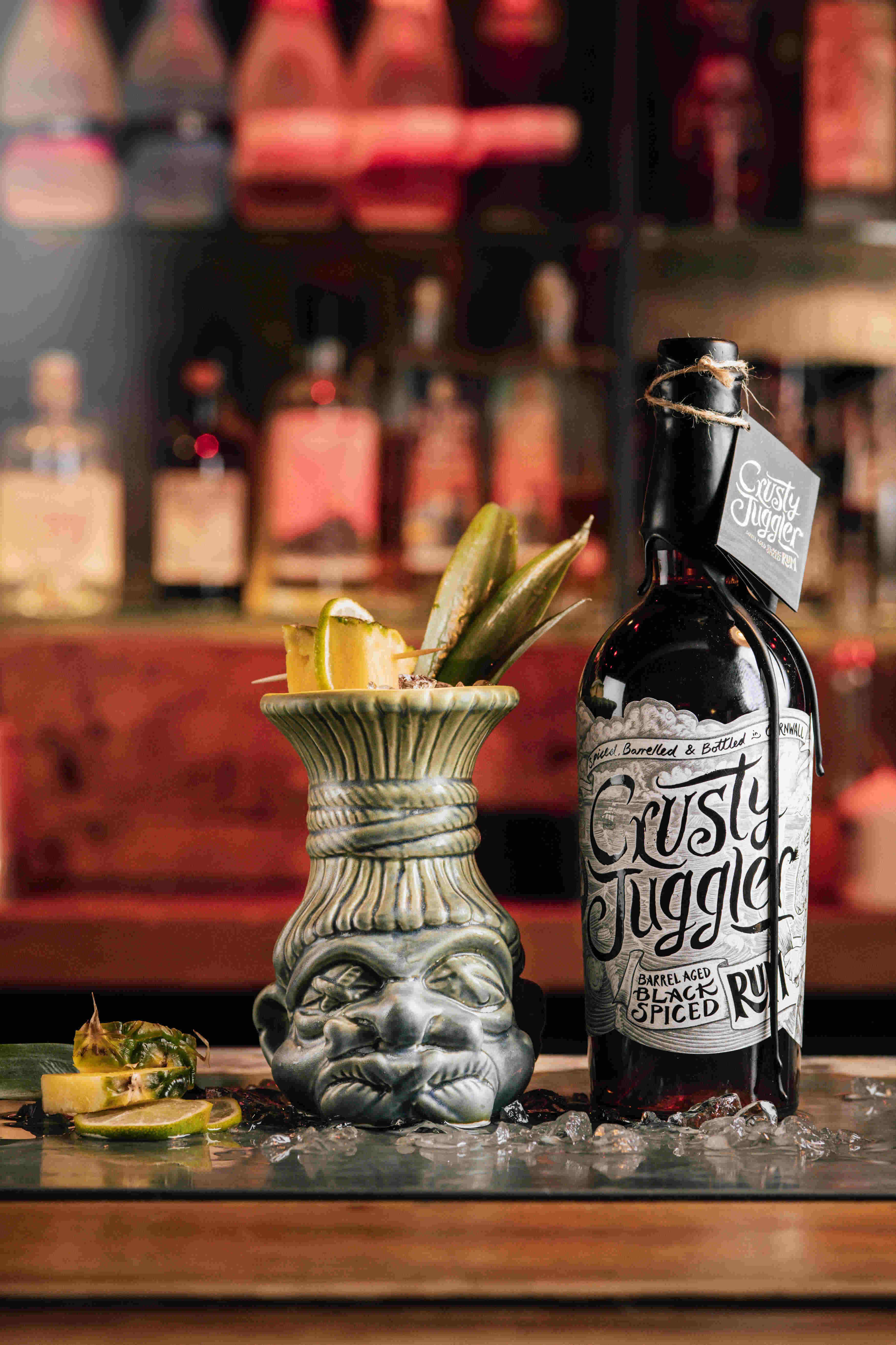 A 70cl bottle of Crusty Juggler Barrel Aged Black Spiced Rum stands on a bar beside a Pineapple Mojito cocktail in a tiki mug. Blurred bottles can be seen in the background.