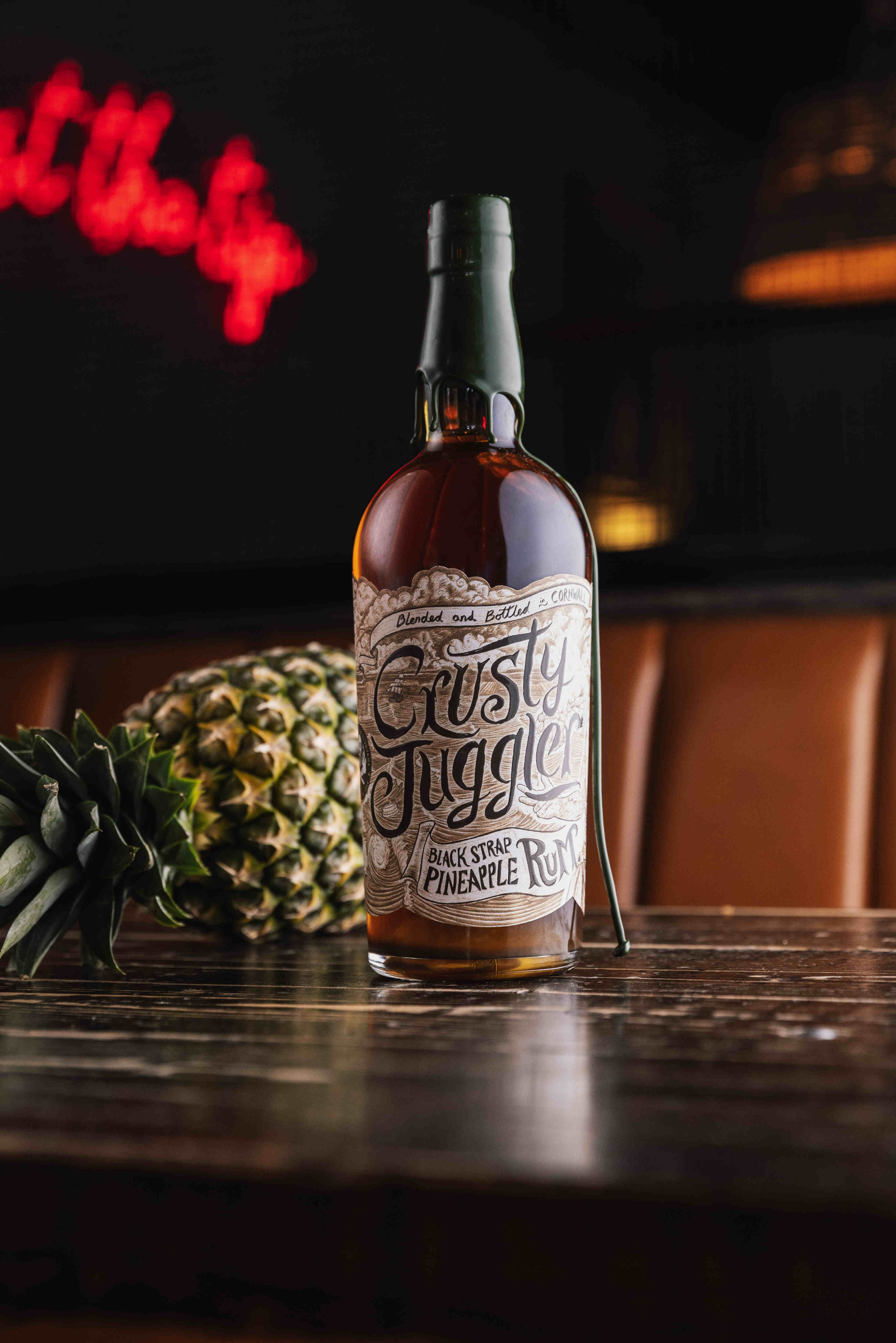 A 70cl bottle of Crusty Juggler Black Strap Pineapple Rum stands on a rustic wooden table beside a pineapple, with warm lighting and a blurred neon sign in the background.