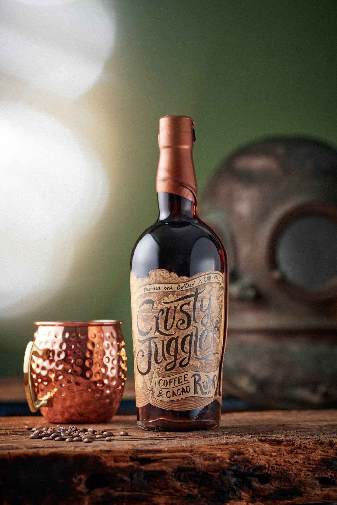 A 70cl bottle of Crusty Juggler Coffee and Cacao Rum stands beside a large Copper Mule Mug on a rustic wooden table against a green background. A vintage scuba diver helmet can be seen in the background.