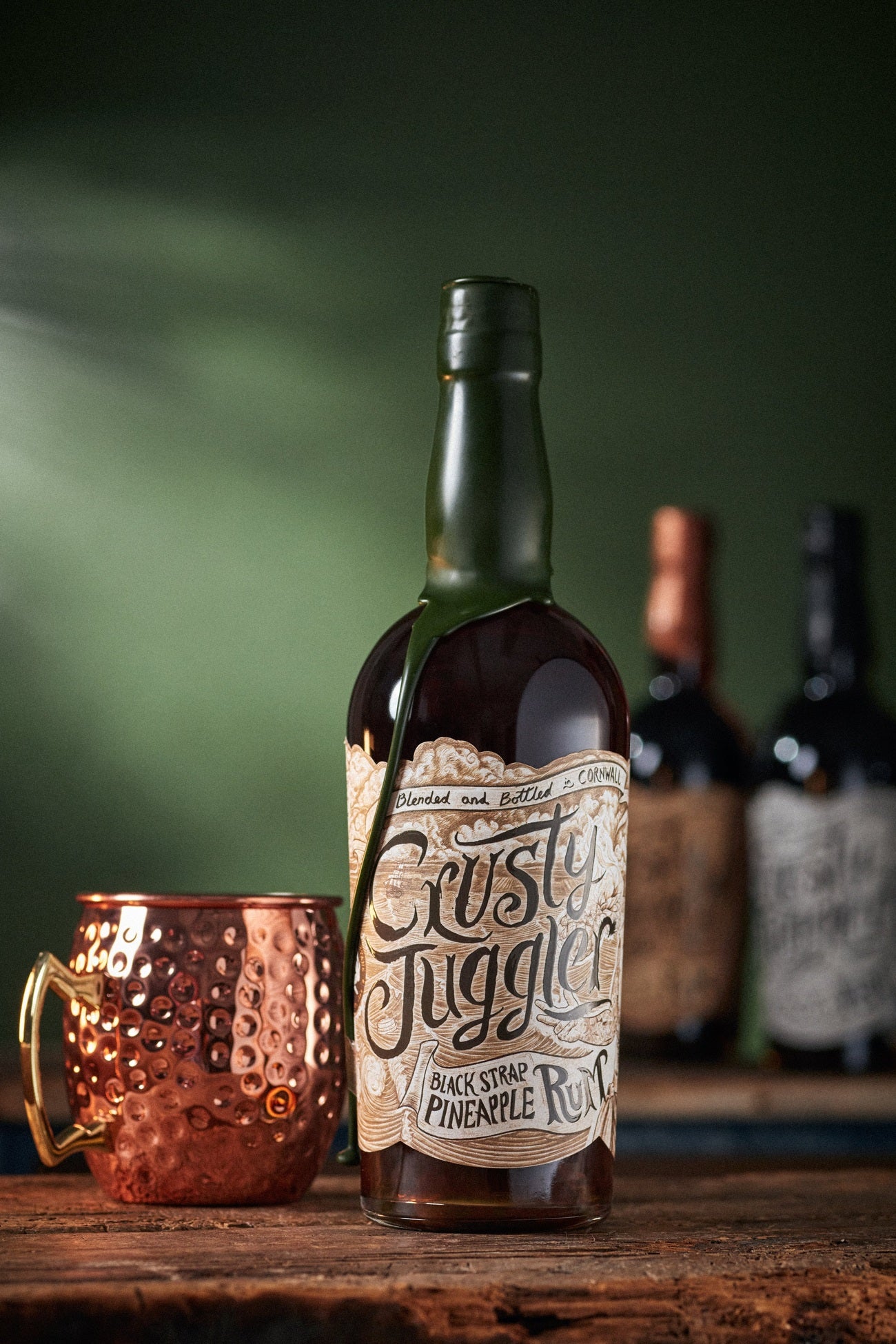 A 70cl bottle of Crusty Juggler Black Strap Pineapple Rum stands beside a large Copper Mule Mug on a rustic wooden table against a green background. Blurred bottles can be seen in the background.