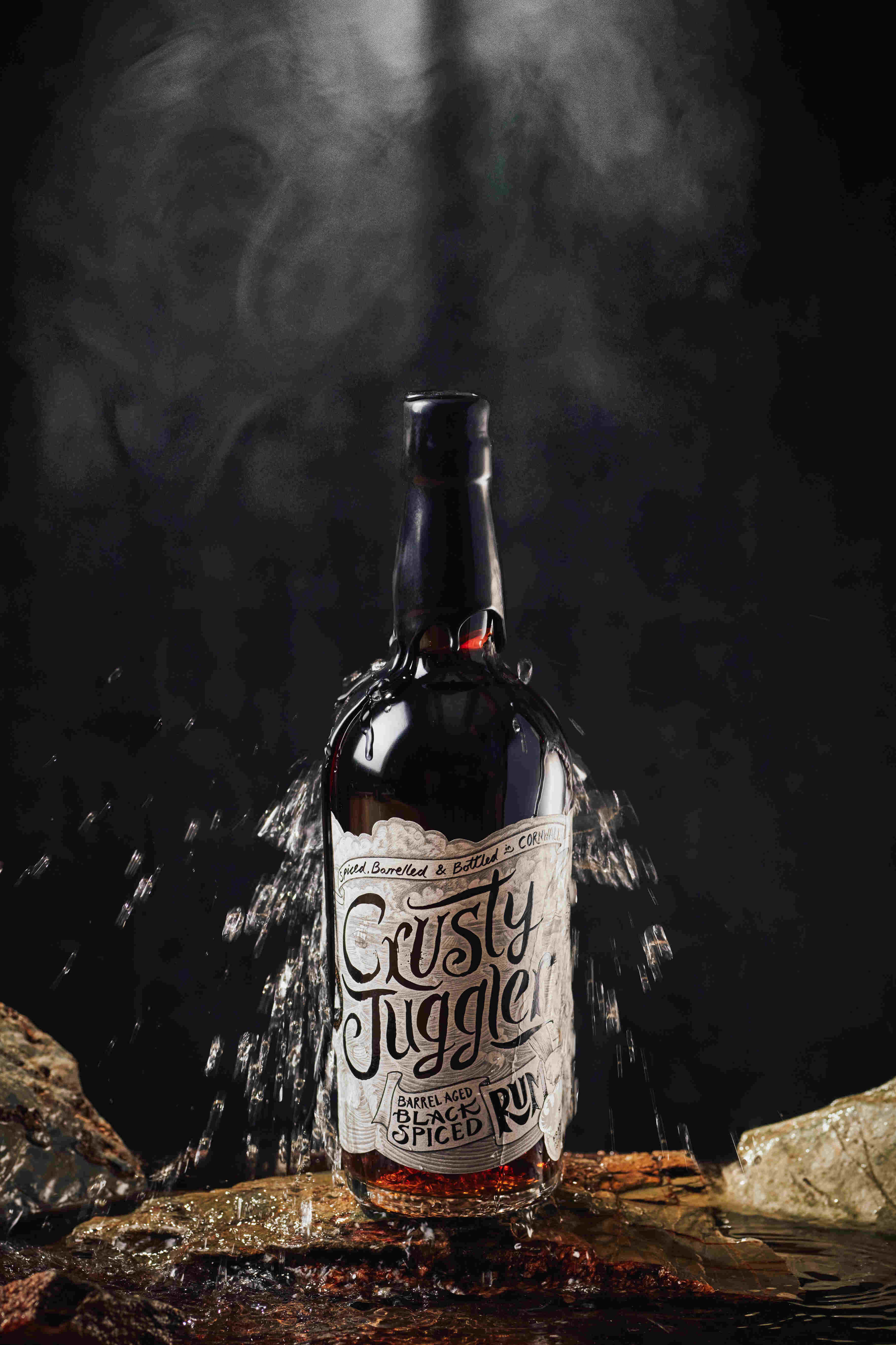 A 70cl bottle of Crusty Juggler Barrel Aged Black Spiced Rum stands on a stone surface against a black background. Water splashes over the bottle from above.