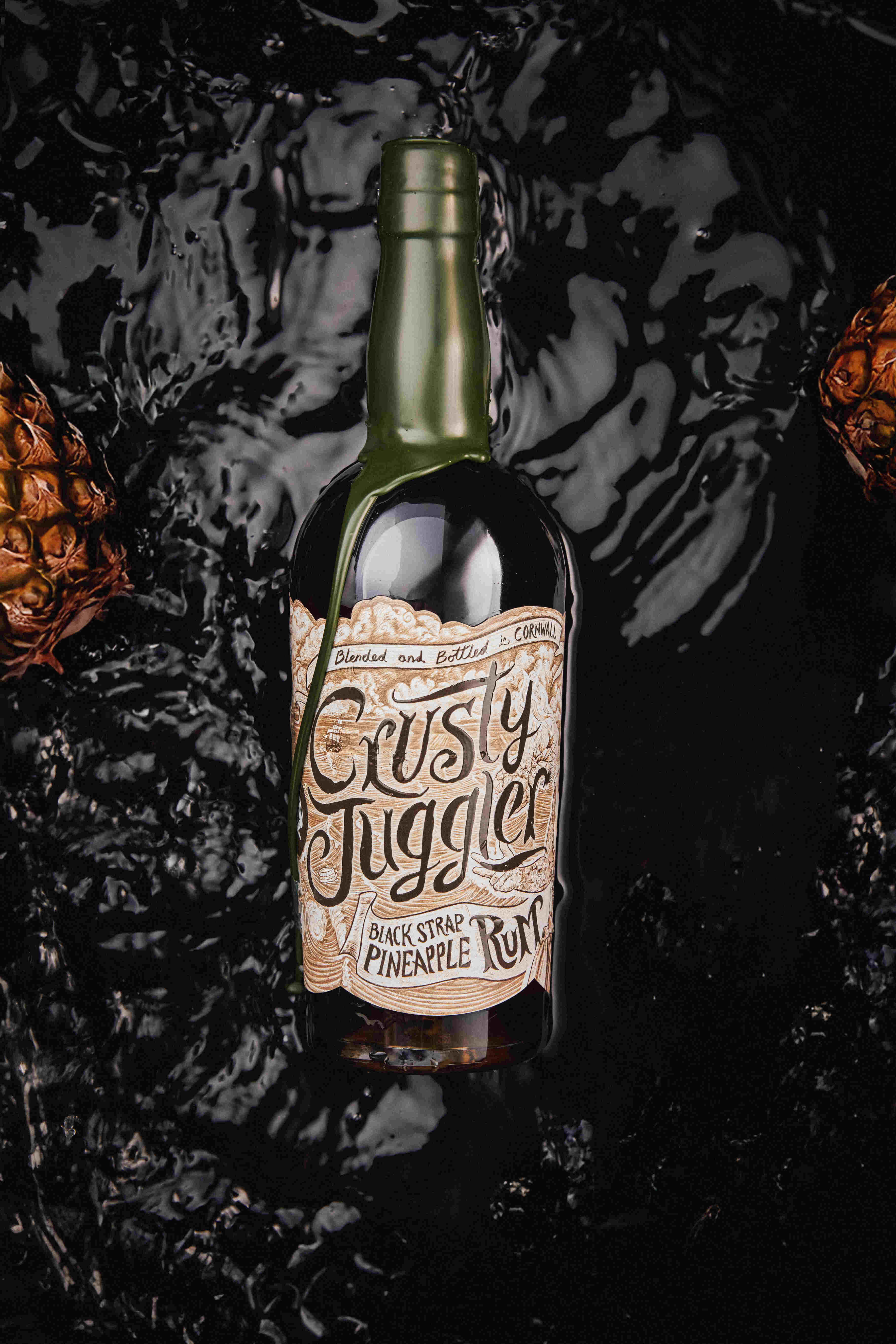 A 70cl bottle of Crusty Juggler Black Strap Pineapple Rum lies on a textured black surface, framed by partially visible pineapples.