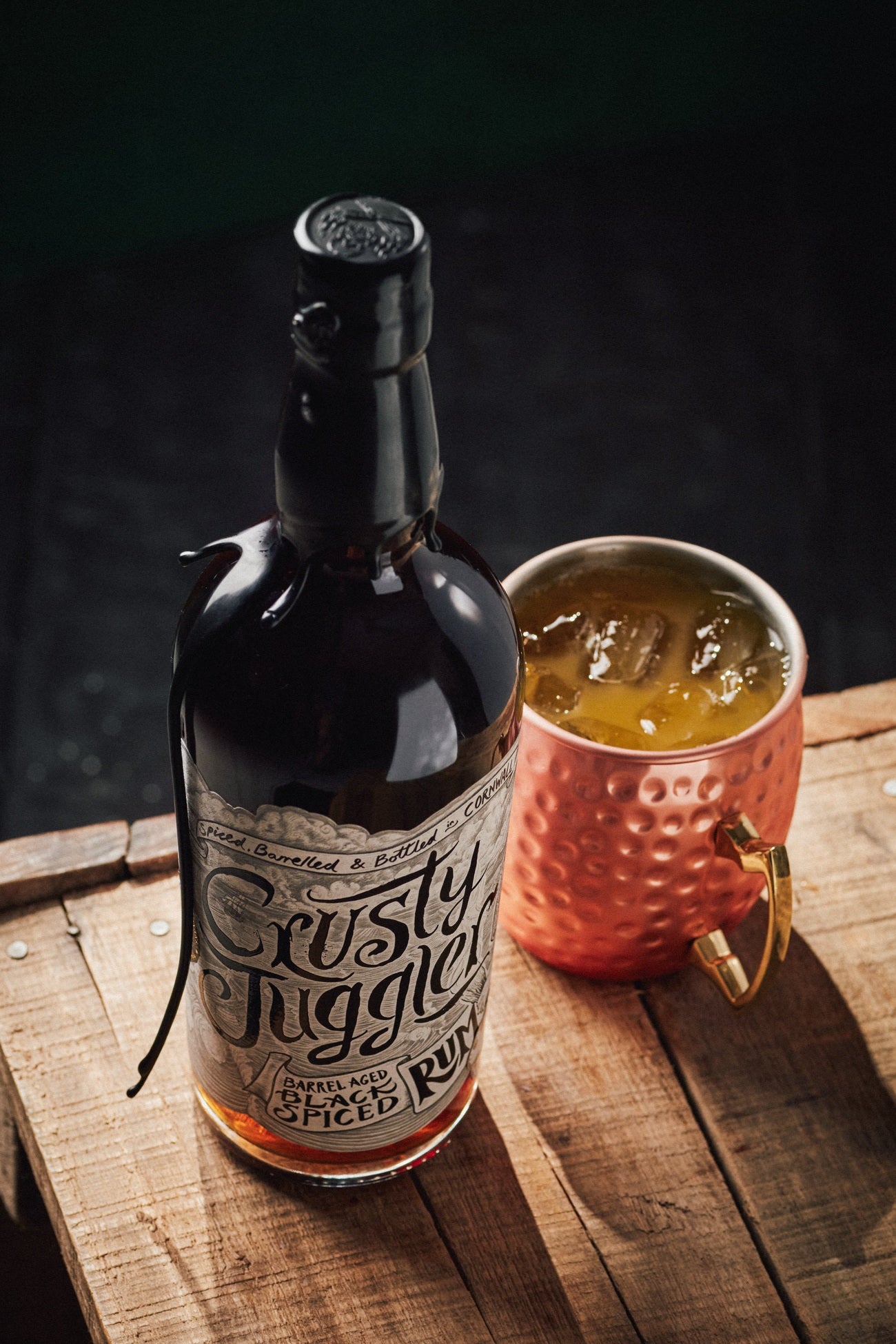 A 70cl bottle of Crusty Juggler Barrel Aged Black Spiced Rum stands beside a large Copper Mule Mug on a rustic wooden table against a black background. The mug contains a Juggler's Revenge cocktail and ice cubes.