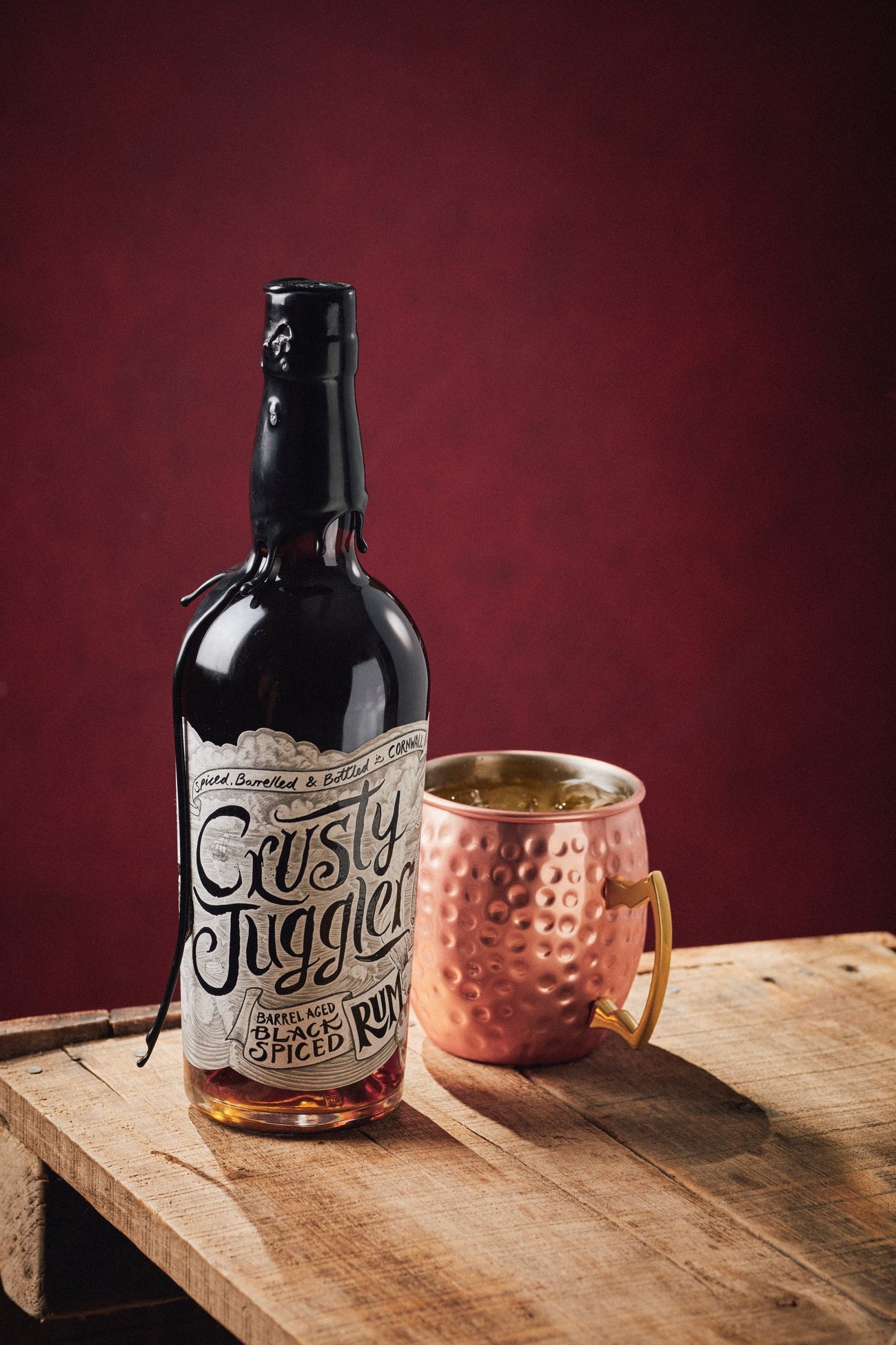 A 5cl bottle of Crusty Juggler Barrel Aged Black Spiced Rum stands beside a large Copper Mule Mug on a rustic wooden table against a red background.