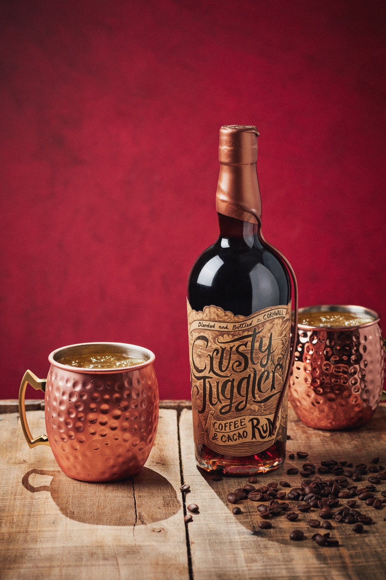 A 70cl bottle of Crusty Juggler Coffee and Cacao Rum stands next to two large Copper Mule Mugs surrounded by coffee beans. They sit on a rustic wooden table against a red background.