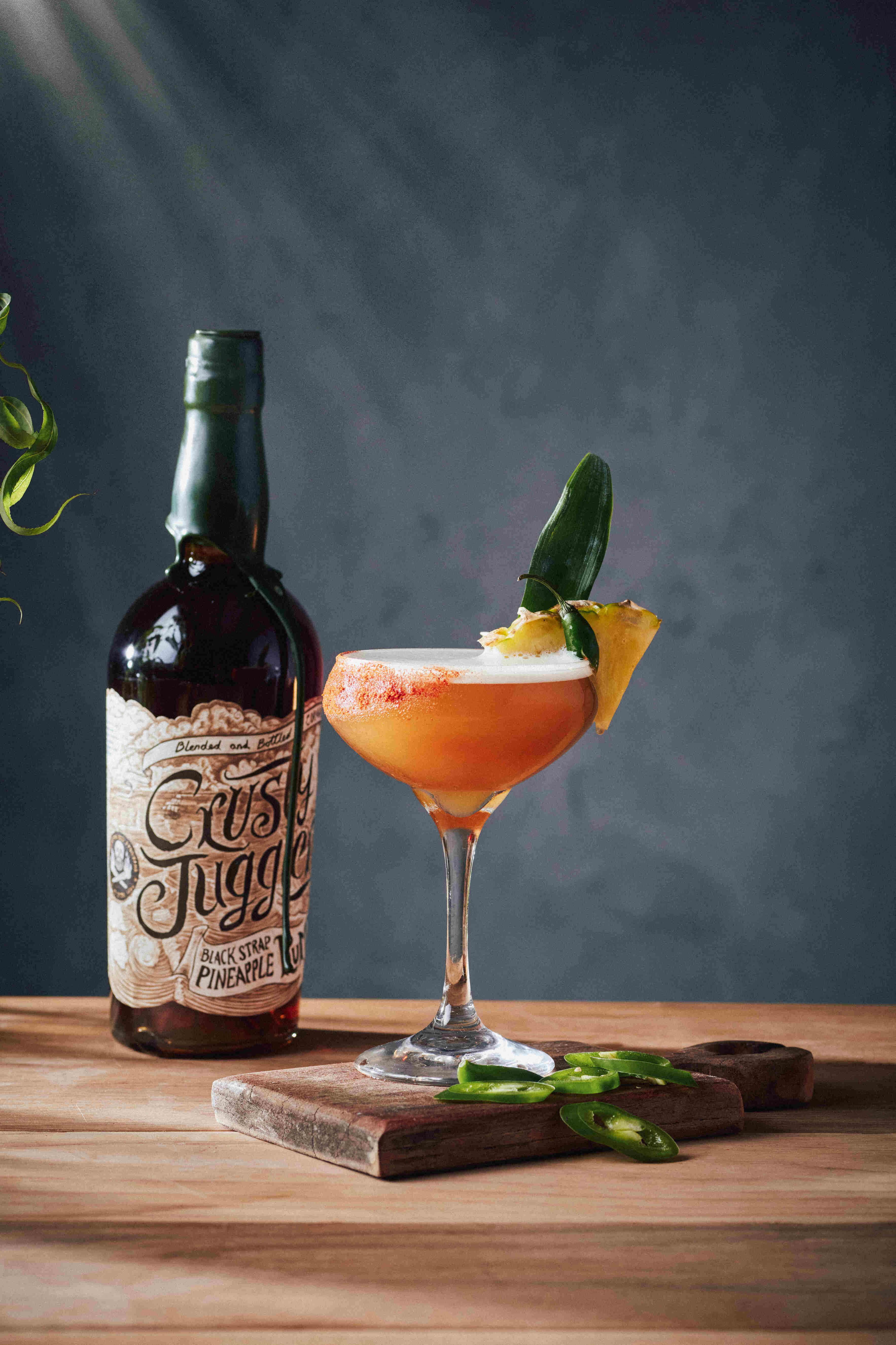 A Pineapple Jalapeno Daiquiri cocktail in a coupe glass stands on a wooden board beside a 70cl bottle of Crusty Juggler Black Strap Pineapple Rum next to some green chilli slices.