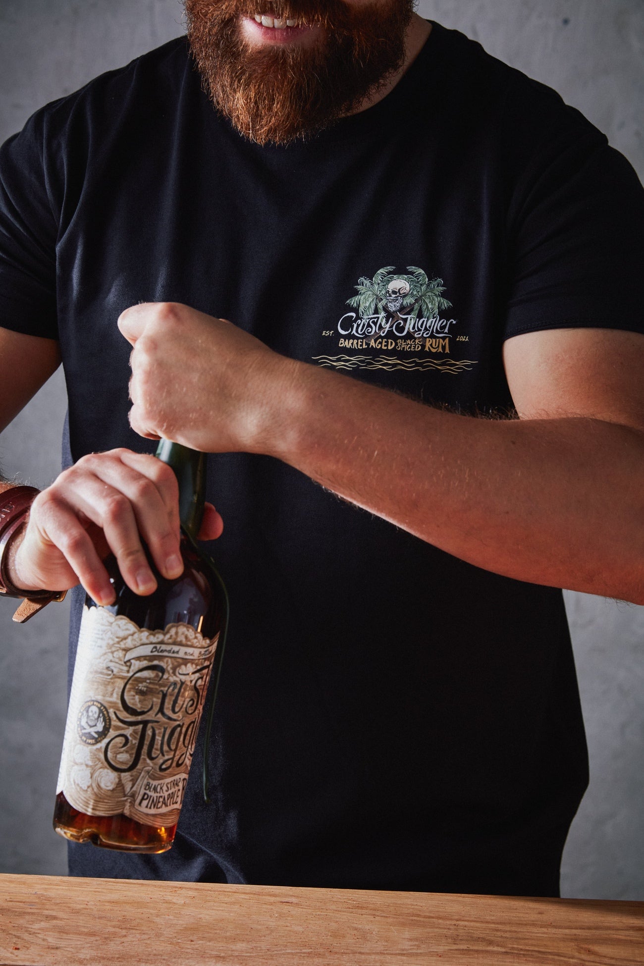 A bearded man holding a bottle of Crusty Juggler Black Strap Pineapple Rum, wears the Crusty Chill Out T-Shirt. The T-shirt is black and features a graphic on the left chest that says "Crusty Juggler Barrel Aged Black Spiced Rum" next to an illustration of palm trees and a skull.