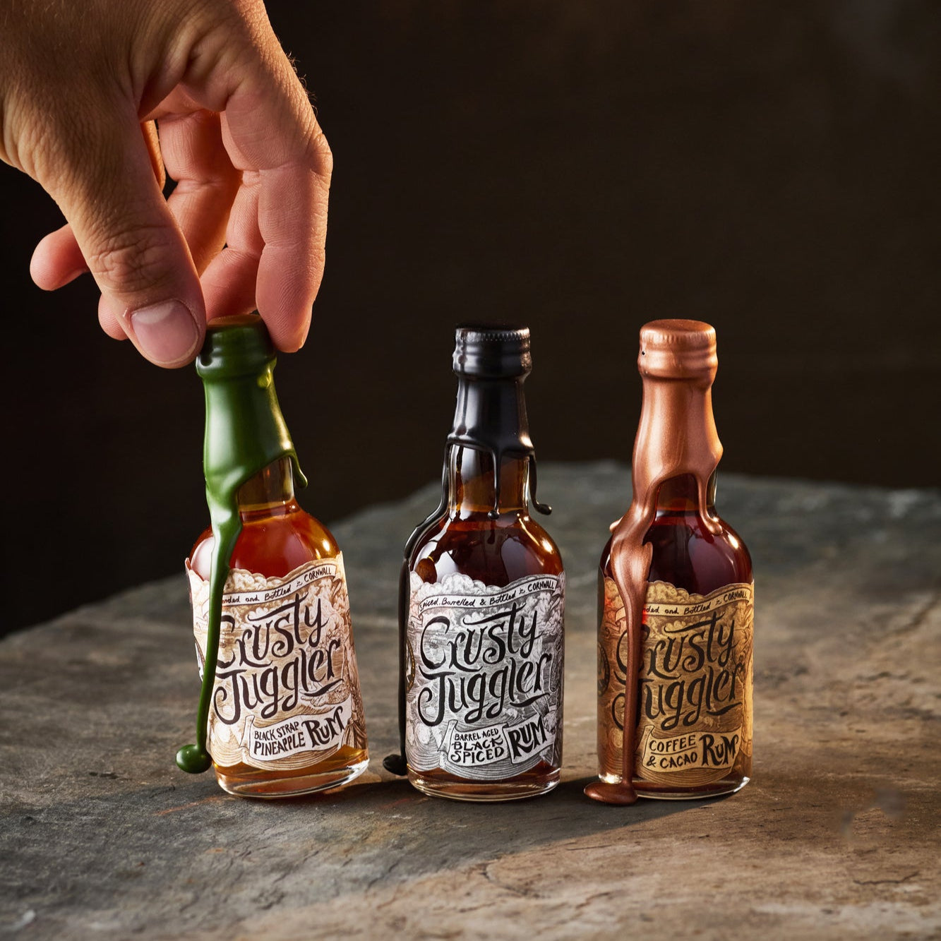 A hand reaches for the Crusty Juggler Miniature Rum Selection, featuring the Crusty Juggler Barrel Aged Black Spiced Rum, the Crusty Juggler Black Strap Pineapple Rum, and the Crusty Juggler Coffee and Cacao Rum. The bottles stand on a rustic stone table against a black background.