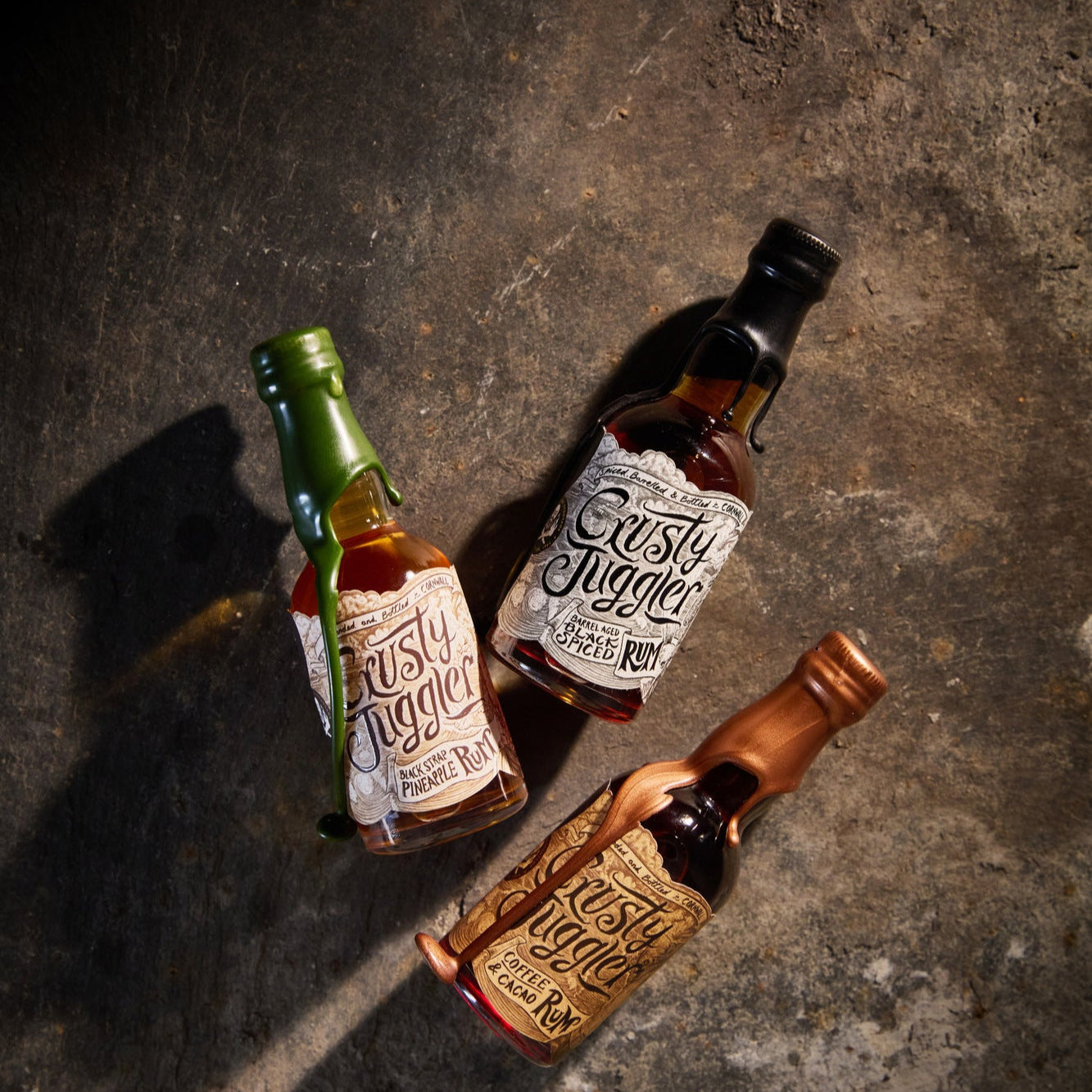 The Crusty Juggler Miniature Rum Selection, featuring the Crusty Juggler Barrel Aged Black Spiced Rum, the Crusty Juggler Black Strap Pineapple Rum, and the Crusty Juggler Coffee and Cacao Rum, are arranged on a rustic stone surface.