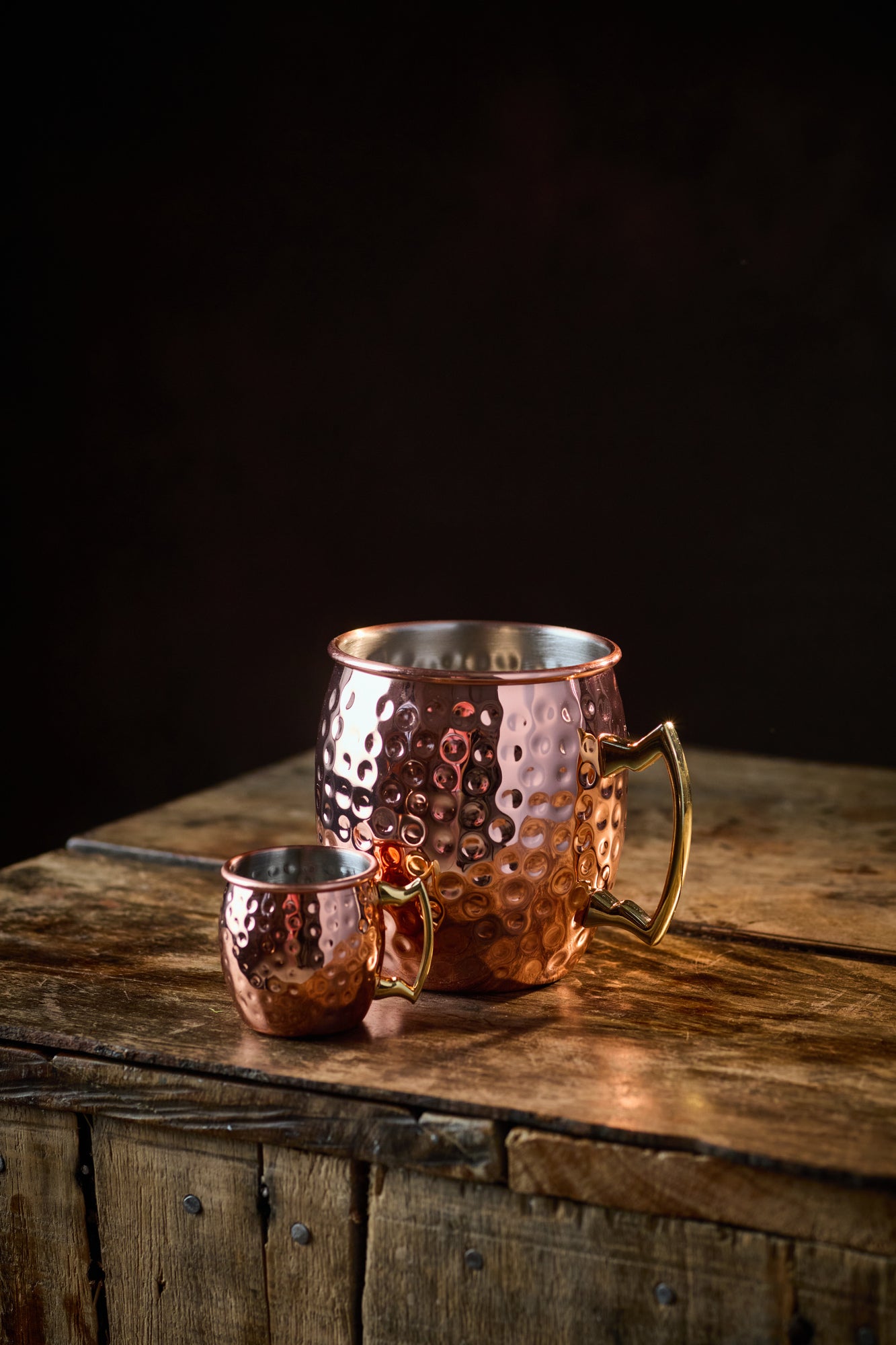 A large and a small Copper Mule Mug, featuring a textured copper-effect finish and a golden handle, sit on a rustic wooden table against a black background.