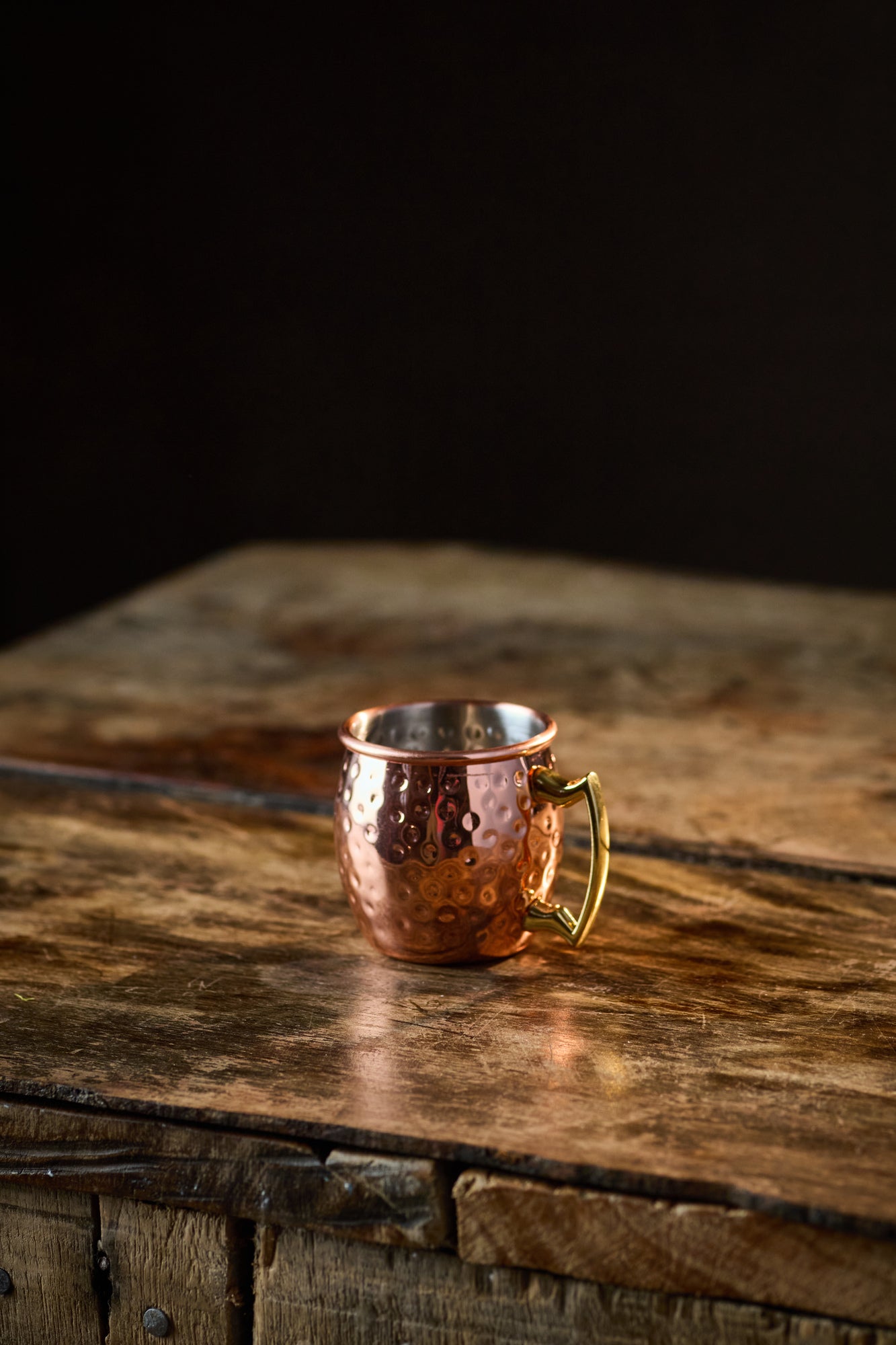 A small Copper Mule Mug, featuring a textured copper-effect finish and a golden handle, sits on a rustic wooden table against a black background.