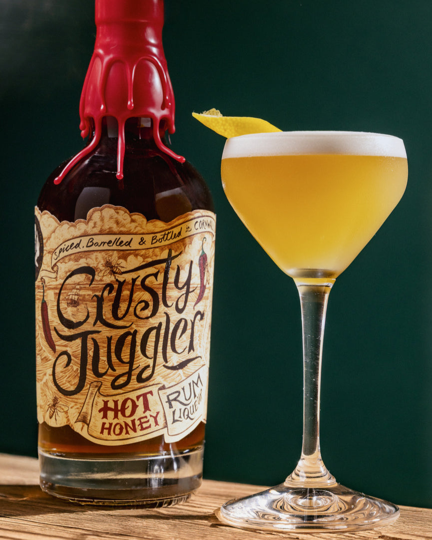 Bottle of 'Crusty Juggler' hot honey rum with a cocktail glass on a wooden surface.