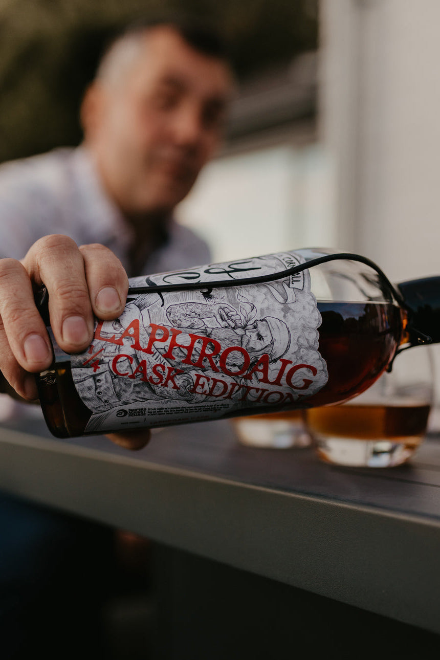 Person holding a bottle of 'Laphroaig Cask Edition' with a glass of liquid in the background.