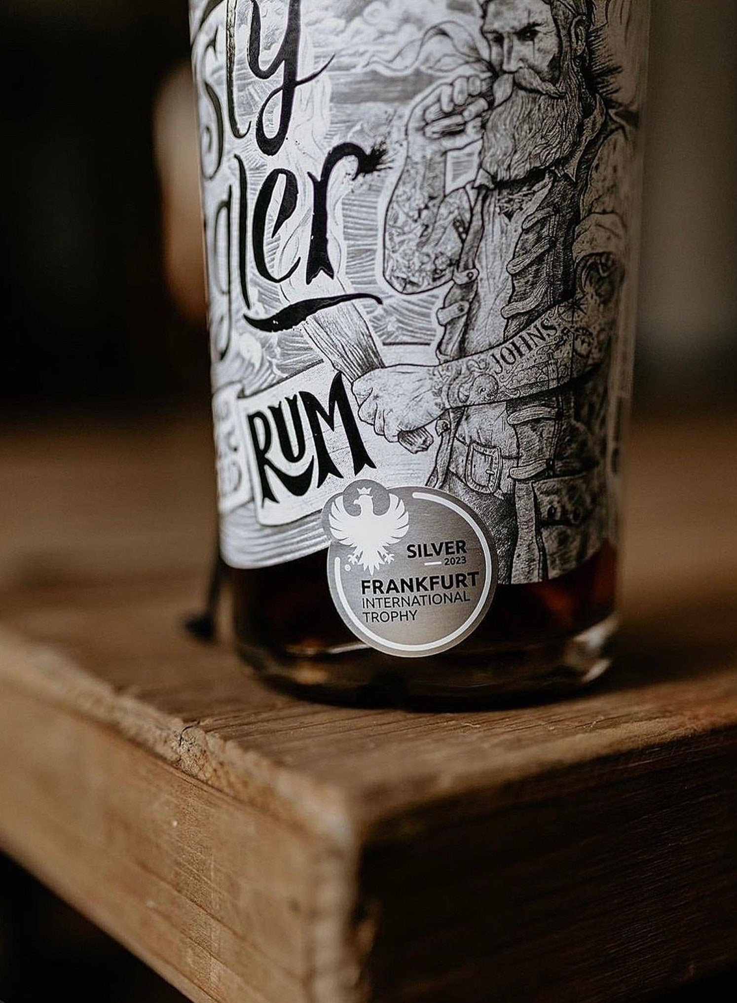 A close-up of a Crusty Juggler Barrel Aged Black Spiced Rum bottle. The label features intricate black-and-white artwork and a silver Frankfurt International Trophy award sticker. The bottle stands on a rustic wooden table.