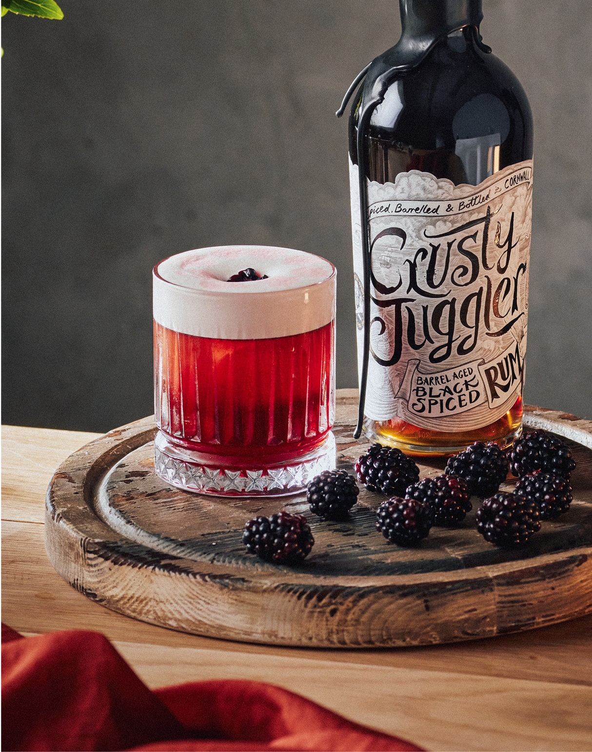 A Crusty Bramble cocktail in a rocks glass and garnished with a blackberry sits on a rustic wooden board surrounded by more blackberries. It sits next to a 70cl bottle of Crusty Juggler Barrel Aged Black Spiced Rum.