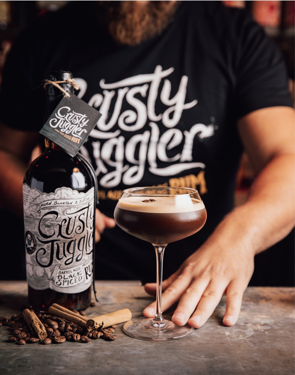 A bearded man wearing the Crusty Classic Black Tee stands behind a bar. A bottle of Crusty Juggler Barrel Aged Black Spiced Rum and an Espresso Crustini cocktail in a coupe glass stand on the counter, next to some cinnamon sticks.