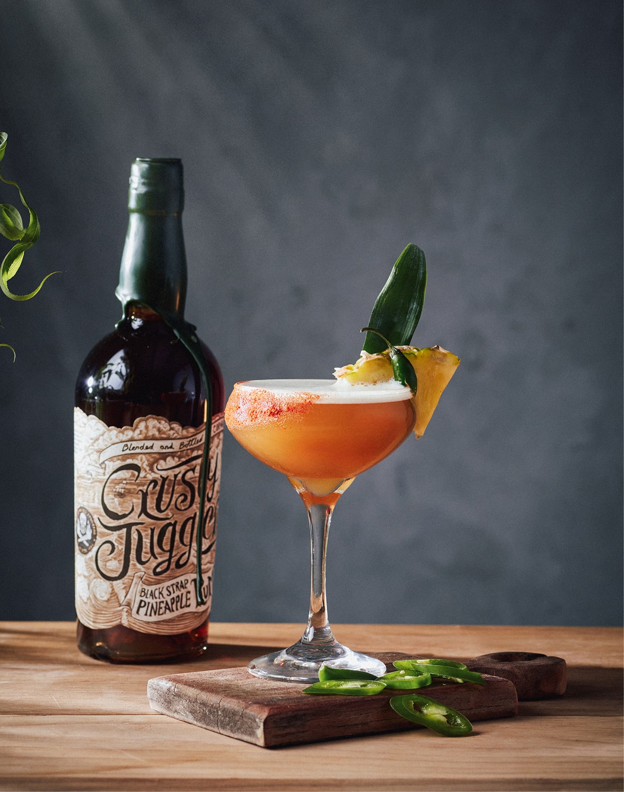 A Pineapple Jalapeno Daiquiri cocktail in a coupe glass stands on a wooden board beside a 70cl bottle of Crusty Juggler Black Strap Pineapple Rum next to some green chilli slices.