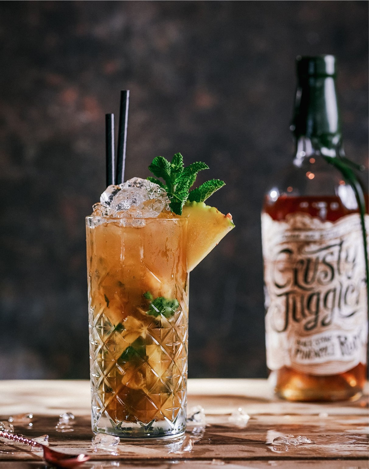 A Pineapple Mojito cocktail in a tall glass garnished with pineapple and mint, stands on a wooden table next to a 70cl bottle of Crusty Juggler Black Strap Pineapple Rum against a textured black background.