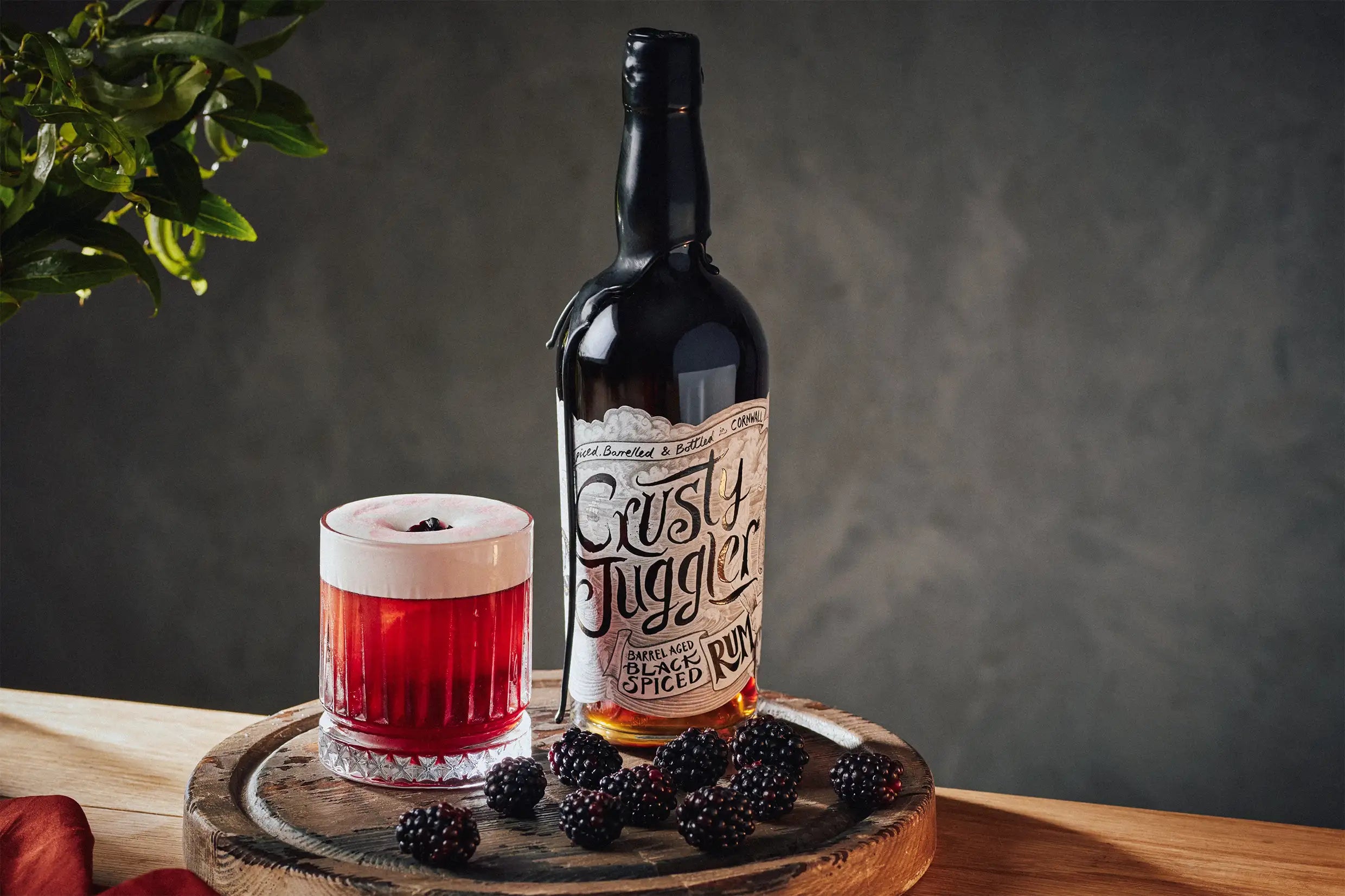 Bottle of 'Crusty Juggler' rum with a glass of red cocktail and blackberries on a wooden surface.