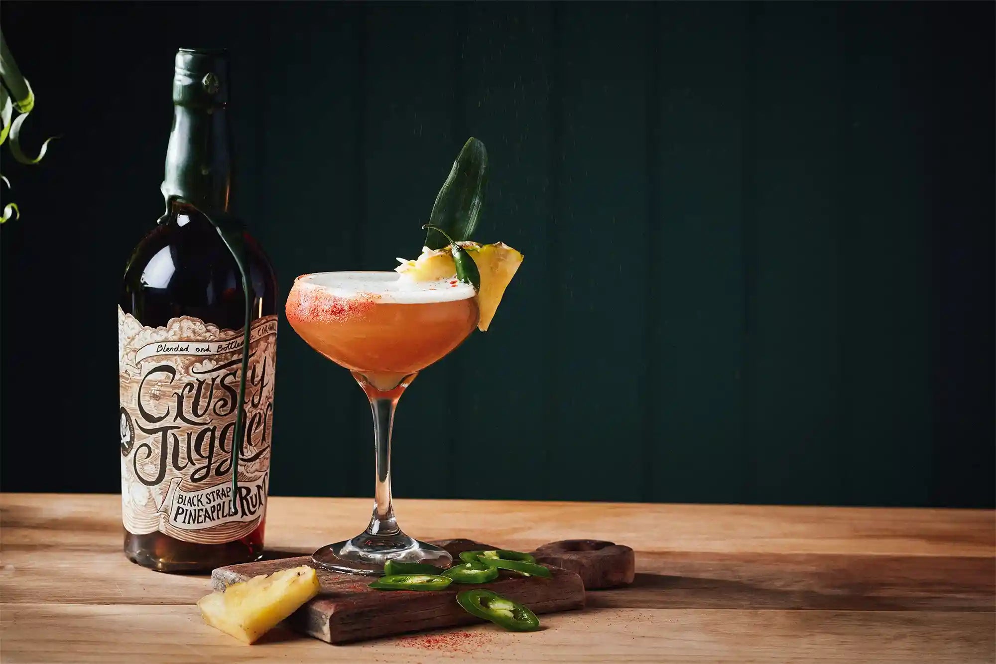Cocktail in a coupe glass with garnishes on a wooden surface, accompanied by a bottle.