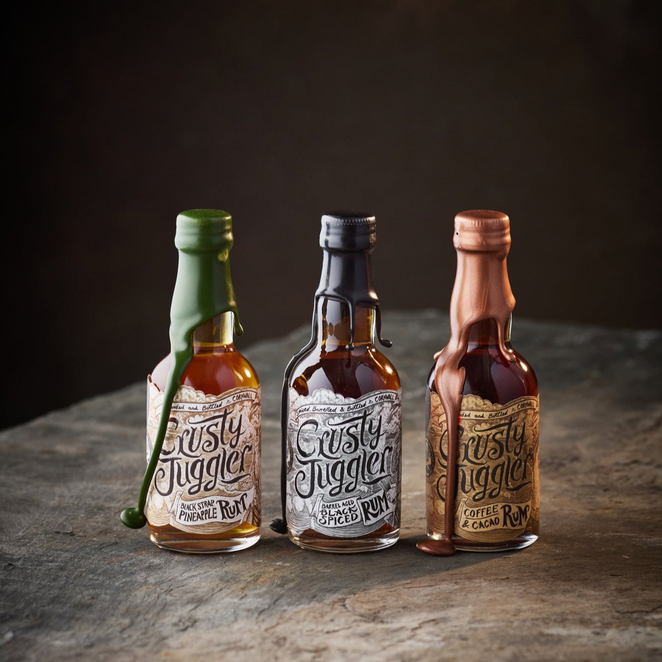 The Crusty Juggler Miniature Rum Selection, featuring the Crusty Juggler Barrel Aged Black Spiced Rum, the Crusty Juggler Black Strap Pineapple Rum, and the Crusty Juggler Coffee and Cacao Rum, stand on rustic stone table against a black background.