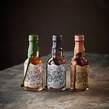 The Crusty Juggler Miniature Rum Selection, featuring the Crusty Juggler Barrel Aged Black Spiced Rum, the Crusty Juggler Black Strap Pineapple Rum, and the Crusty Juggler Coffee and Cacao Rum, stand on rustic stone table against a black background.