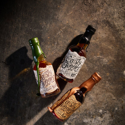 The Crusty Juggler Miniature Rum Selection, featuring the Crusty Juggler Barrel Aged Black Spiced Rum, the Crusty Juggler Black Strap Pineapple Rum, and the Crusty Juggler Coffee and Cacao Rum, are arranged on a rustic stone surface.