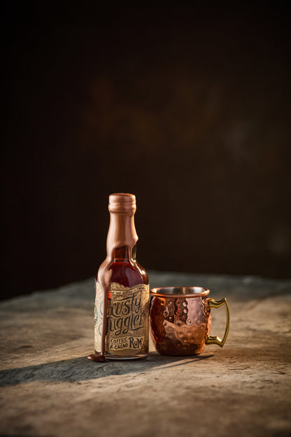 A 5cl bottle of Crusty Juggler Coffee and Cacao Rum stands beside a small Copper Mule Mug on a rustic stone table against a black background.