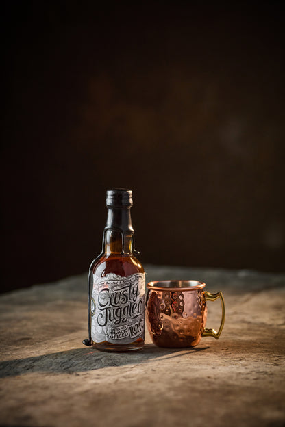 A 5cl bottle of Crusty Juggler Barrel Aged Black Spiced Rum stands beside a small Copper Mule Mug on a rustic stone table against a black background.