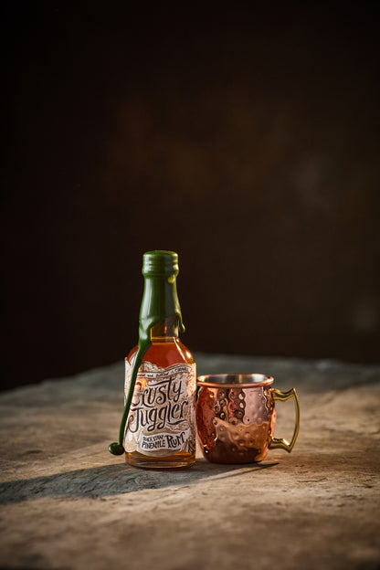 A 5cl bottle of Crusty Juggler Black Strap Pineapple Rum stands beside a small Copper Mule Mug on a rustic stone table against a black background.