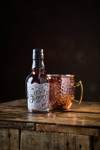 A 20cl bottle of Crusty Juggler Barrel Aged Black Spiced Rum stands beside a large Copper Mule Mug on a rustic wooden table against a black background.