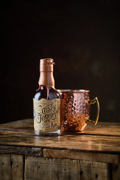 A 20cl bottle of Crusty Juggler Coffee and Cacao Rum stands beside a large Copper Mule Mug on a rustic wooden table against a black background.