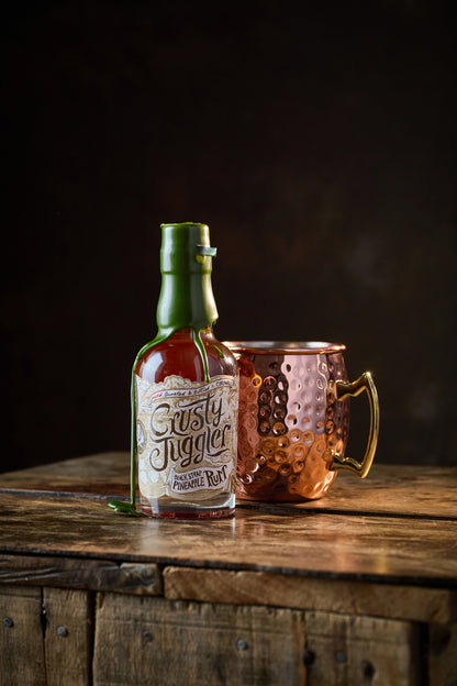 A 20cl bottle of Crusty Juggler Black Strap Pineapple Rum stands beside a large Copper Mule Mug on a rustic wooden table against a black background.