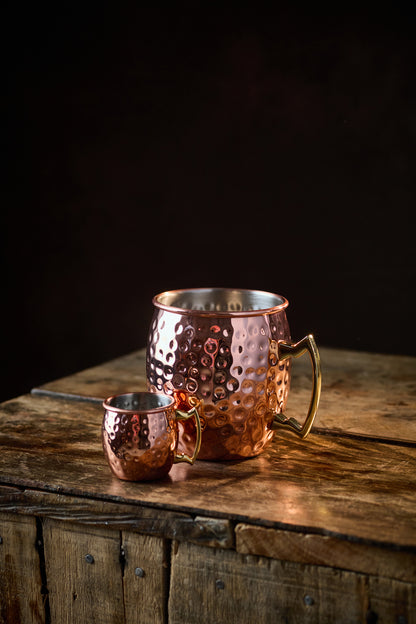 A large and a small Copper Mule Mug, featuring a textured copper-effect finish and a golden handle, sit on a rustic wooden table against a black background.