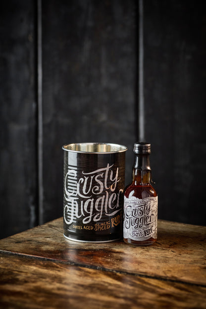 A 5cl bottle of Crusty Juggler Barrel Aged Black Spiced Rum stands beside a Crusty Drinking Can on a rustic wooden table against a textued black background.