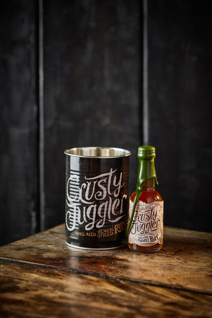 A 5cl bottle of Crusty Juggler Black Strap Pineapple Rum stands beside a Crusty Drinking Can on a rustic wooden table against a textured black background.