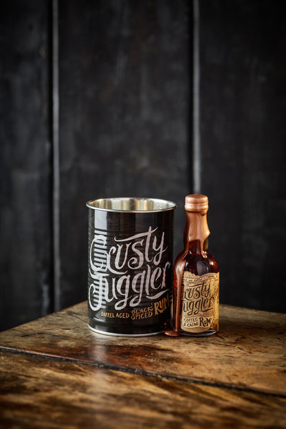 A 5cl bottle of Crusty Juggler Coffee and Cacao Rum stands beside a Crusty Drinking Can on a rustic wooden table against a textured black background.