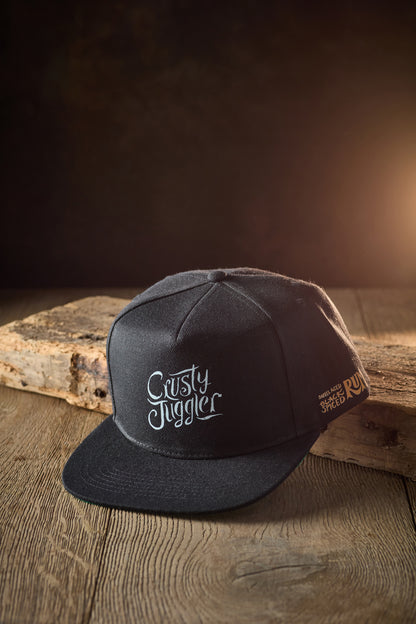 A black Crusty Snapback Cap, featuring the Crusty Juggler logo on the front, sits on a rustic wooden board against a black background with warm lighting. 