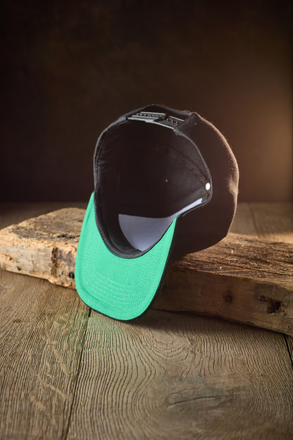 A black Crusty Snapback Cap, featuring a bright green under-brim, sits upside down on a rustic wooden board against a black background with warm lighting.