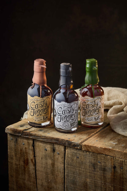 The Crusty Juggler Rum 20cl Selection, featuring the Crusty Juggler Barrel Aged Black Spiced Rum, the Crusty Juggler Black Strap Pineapple Rum, and the Crusty Juggler Coffee and Cacao Rum, stand on a rustic wooden table with a beige cloth against a black background.