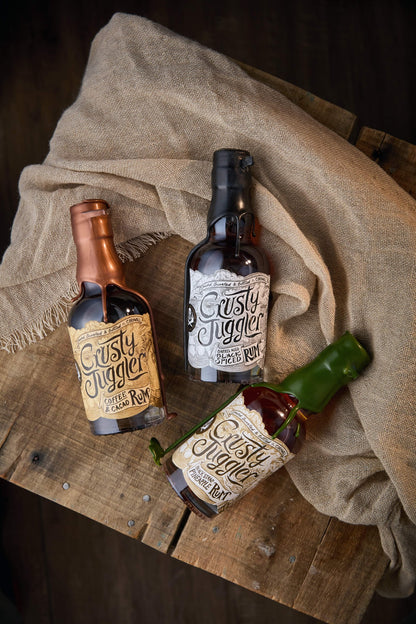 The Crusty Juggler Rum 20cl Selection, featuring the Crusty Juggler Barrel Aged Black Spiced Rum, the Crusty Juggler Black Strap Pineapple Rum, and the Crusty Juggler Coffee and Cacao Rum, are arranged on a rustic wooden table with a beige cloth against a black background.
