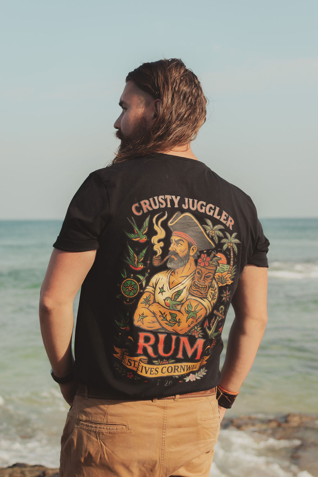 Man wearing a black t-shirt with a colorful graphic and text, standing on a beach.